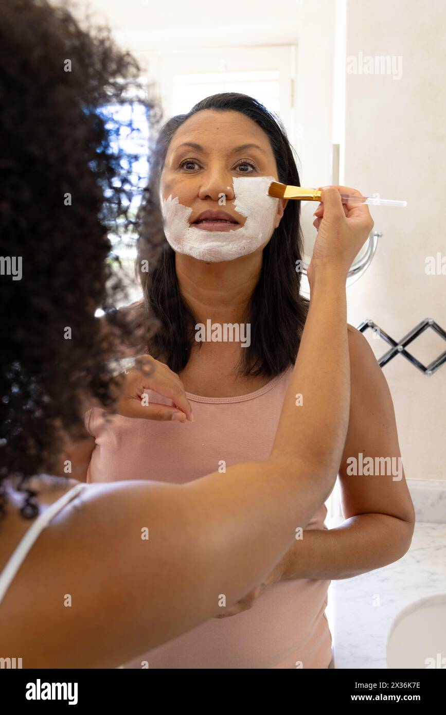 Le donne birazziali mature e le figlie usano maschere facciali, guardando allo specchio Foto Stock