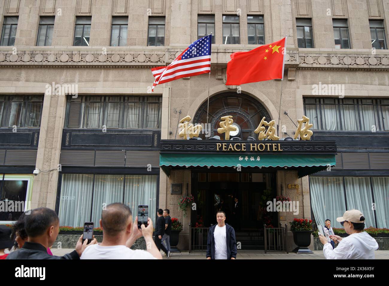 SHANGHAI, CINA - 25 APRILE 2024 - la bandiera rossa a cinque stelle della Cina e la bandiera degli Stati Uniti sono appese sopra l'ingresso del Peace Hotel lungo Nanjing Road Pede Foto Stock