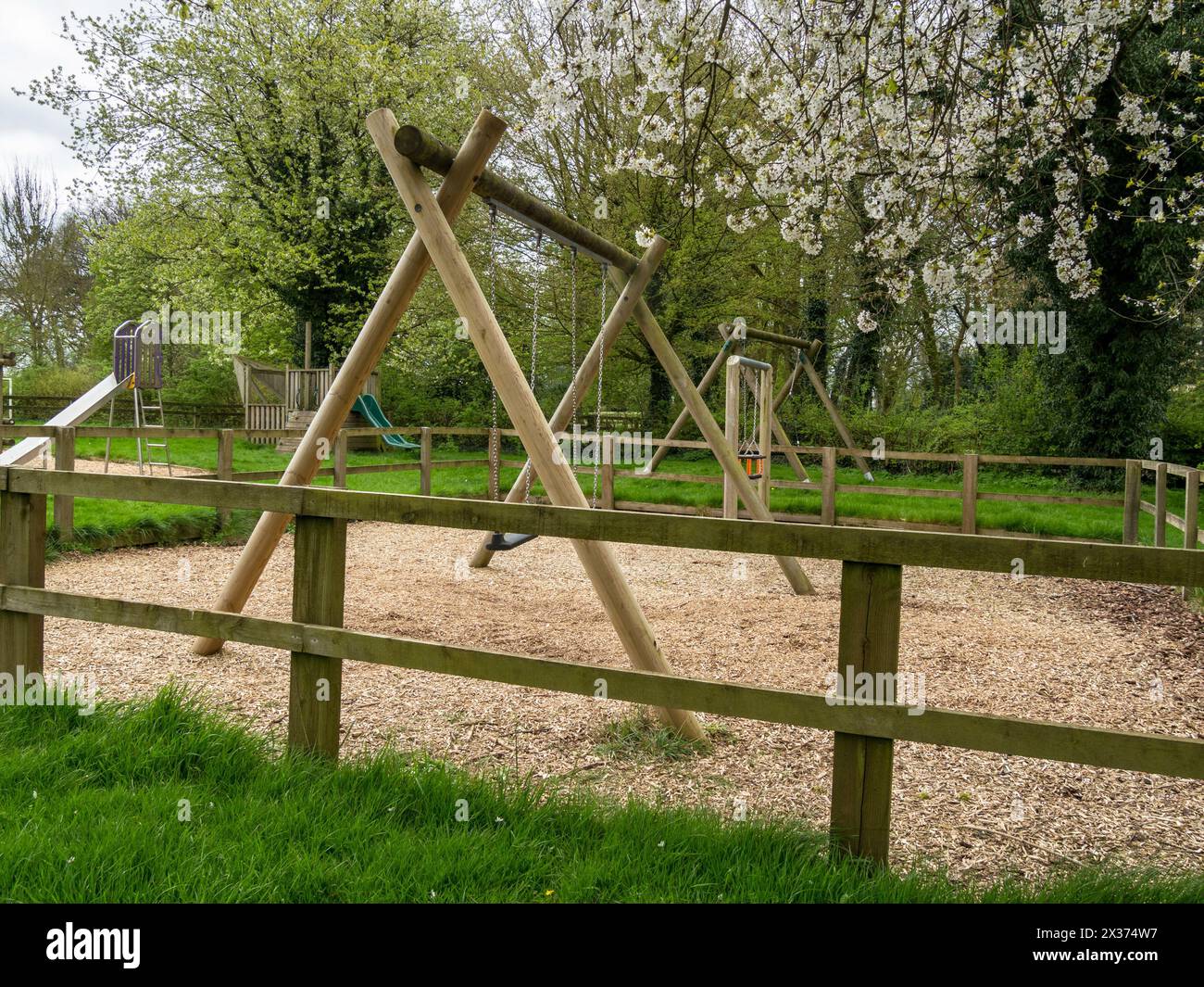 Area giochi per bambini recintata / area ricreativa / parco giochi con altalene e chippate di corteccia sotto, Somerby, Leicestershire, Inghilterra, Regno Unito Foto Stock