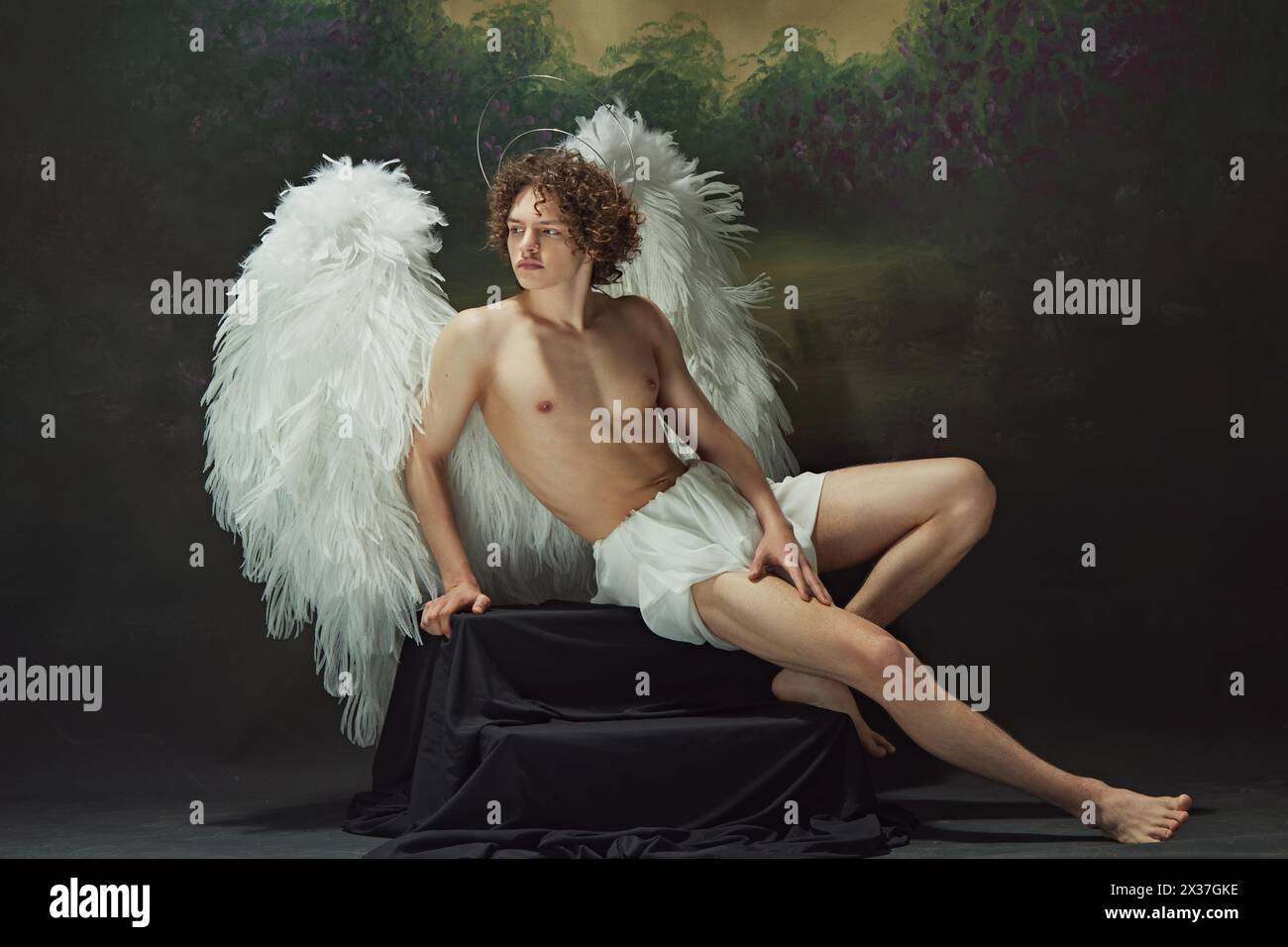 Un uomo affascinante con ricci e ali bianche che si posano irradia un senso di pace come un angelo sullo sfondo di uno studio d'epoca. Foto Stock