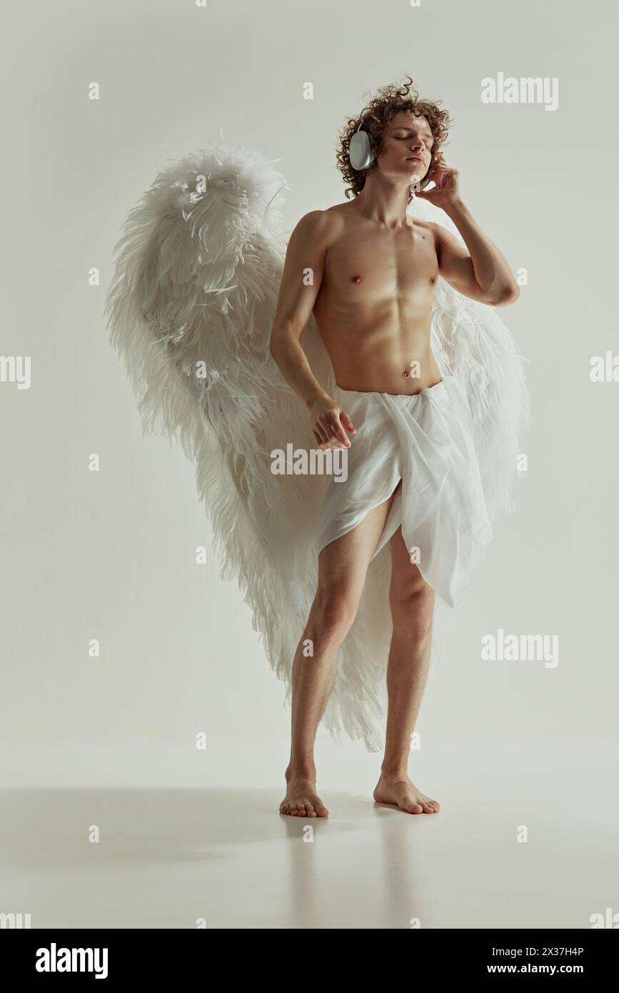 Giovane uomo con grandi ali e angelo che ascolta musica con le moderne cuffie su sfondo bianco da studio. Foto Stock