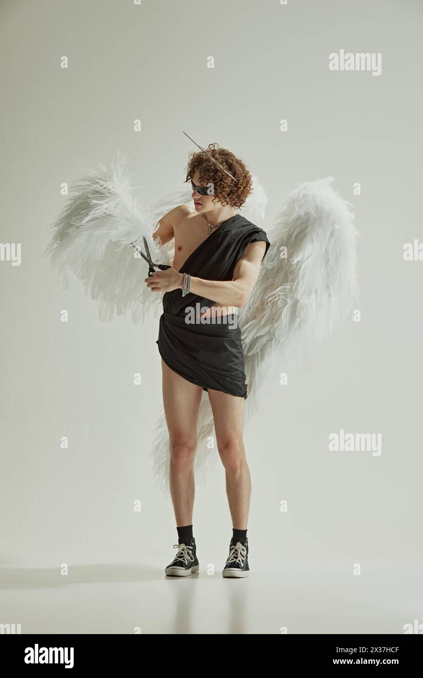 Il giovane si presenta come un personaggio mitologico che taglia le sue grandi ali bianche con forbici su sfondo bianco da studio. Foto Stock