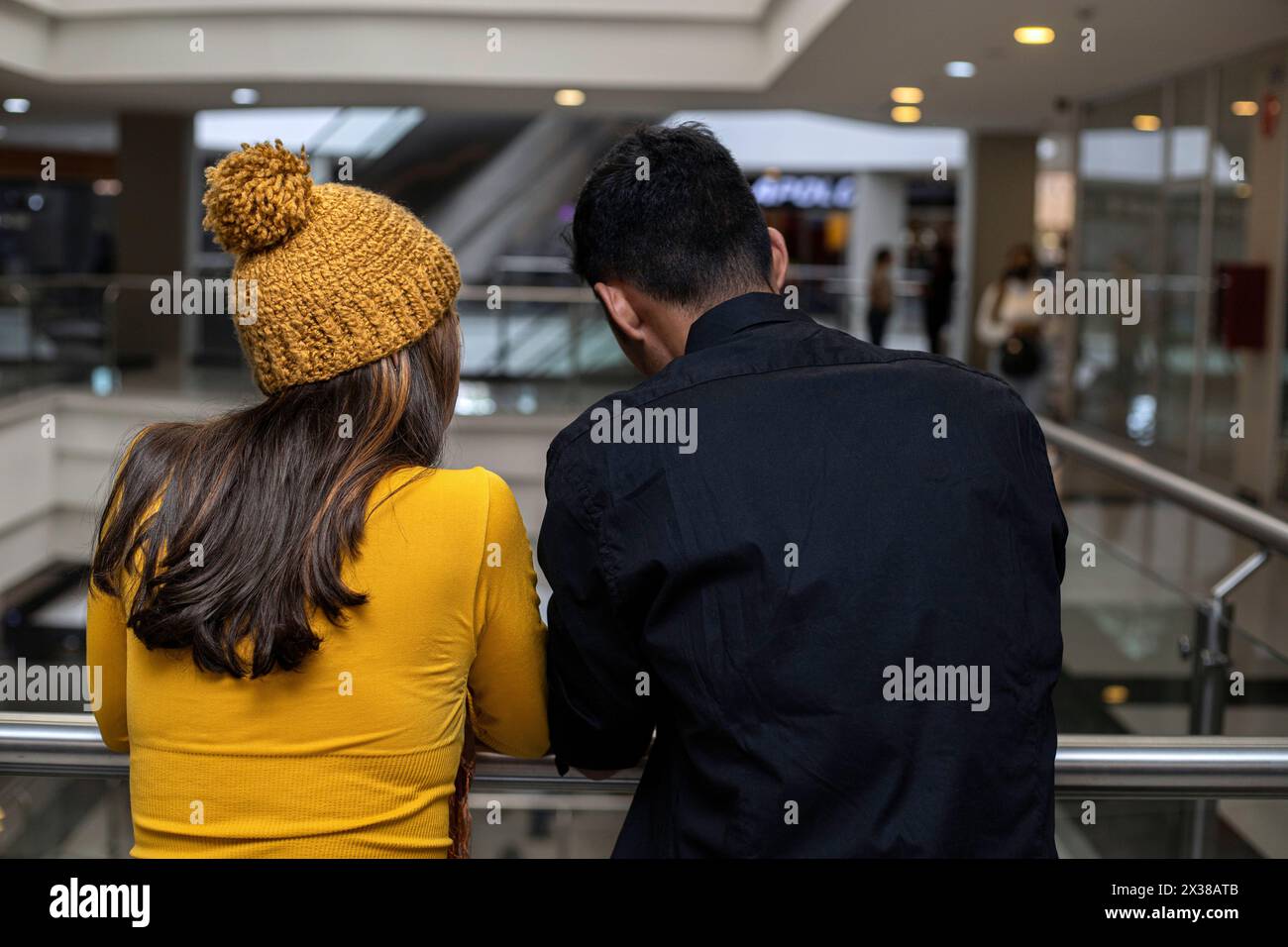 Due latinoamericani sono in piedi con le spalle contro la ringhiera del centro commerciale. Indossa un cappello giallo e un flanella a maniche lunghe, lui è in bla Foto Stock