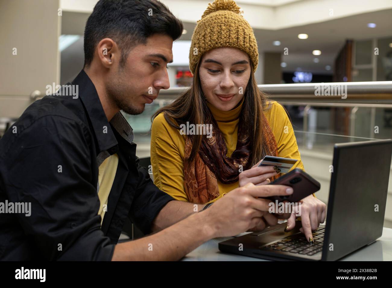 Una giovane coppia latinoamericana tiene in mano una carta di credito e utilizza un computer portatile e un telefono cellulare per effettuare pagamenti via Internet, servizi bancari digitali e sho online Foto Stock