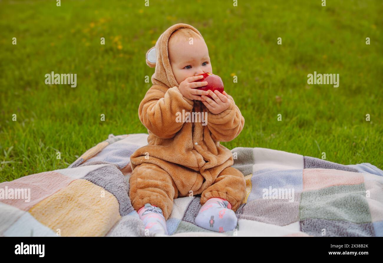 Una bambina con una tuta calda è seduta su un prato verde e mangia una mela Foto Stock