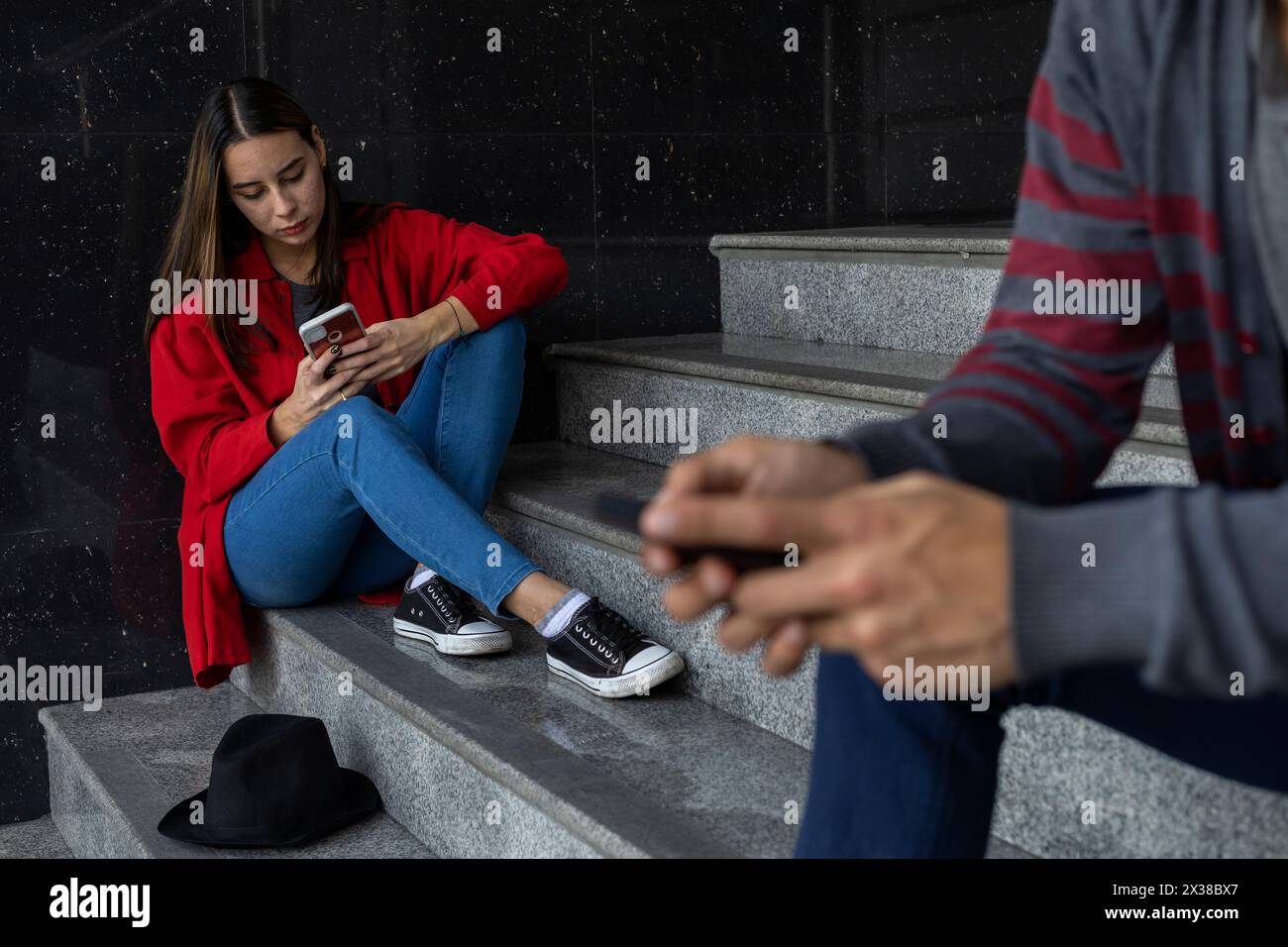 Giovane donna (23) latinoamericana seduta sulle scale a controllare il suo cellulare. Concentrati selettivamente su di lei. Tecnologia e concetto di fratellanza Foto Stock