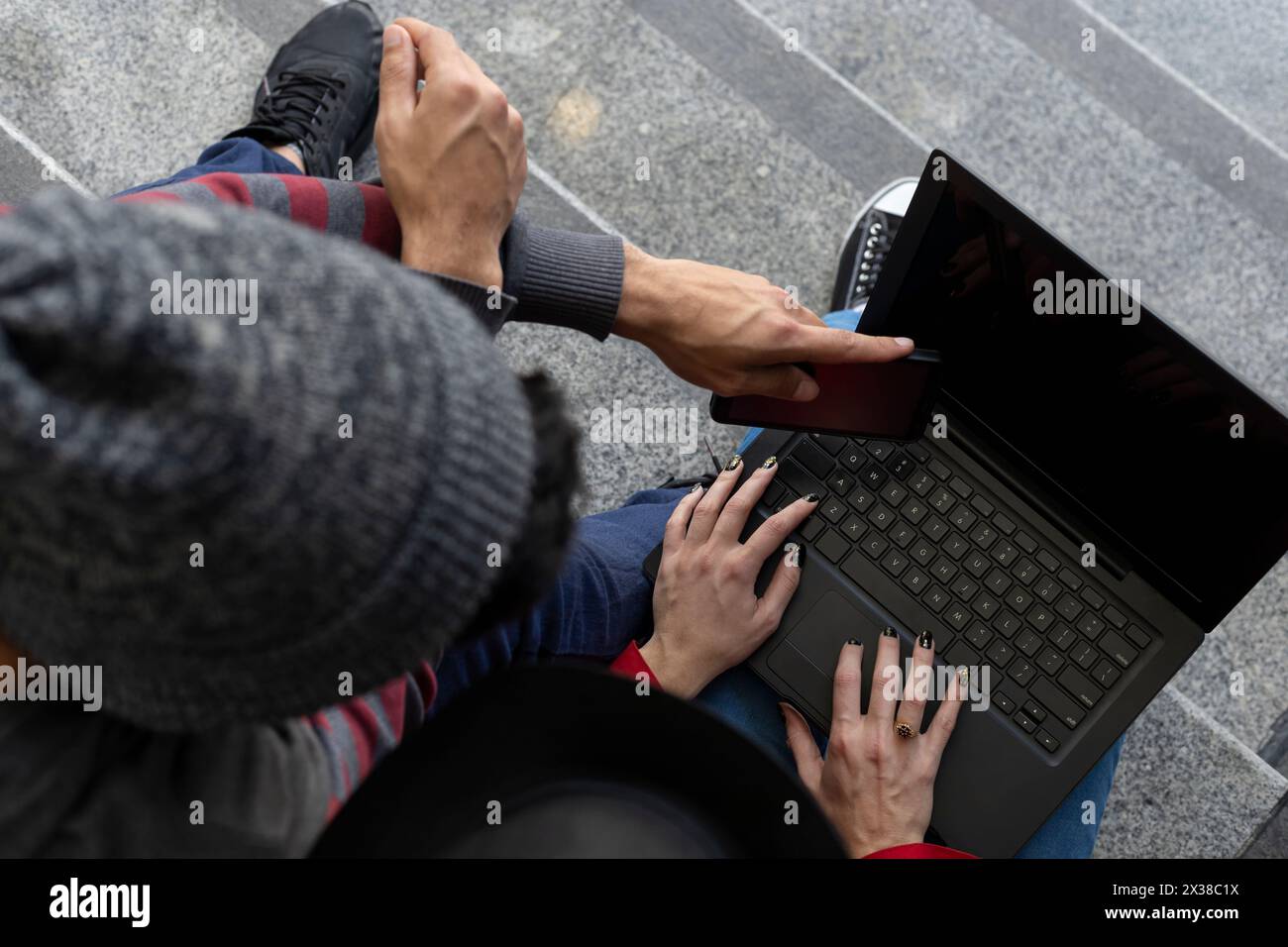 Due giovani e irriconoscibili fratelli freelancer latinoamericani, seduti sulle scale con un computer portatile e un telefono cellulare mentre condividono un po' di tempo di lavoro. Vista da Foto Stock