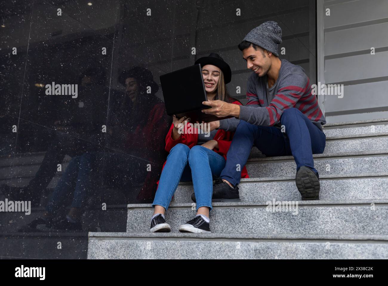 Due giovani fratelli latinoamericani seduti sulle scale con un computer portatile e un telefono cellulare, mentre condividono un po' di tempo di lavoro guardando video e messaggi. Tecnol Foto Stock