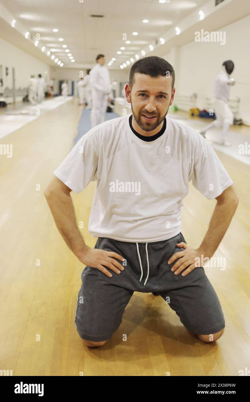 L'uomo siede sul pavimento in palestra per allenarsi con la scherma, altre persone fuori fuoco Foto Stock