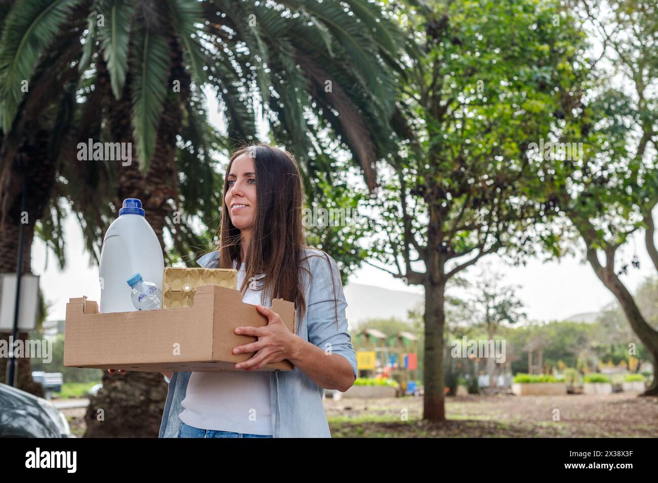 Una donna tiene in mano una scatola di cartone con la spazzatura per riciclare e gettare i bidoni della spazzatura. Sorride ed è felice Foto Stock