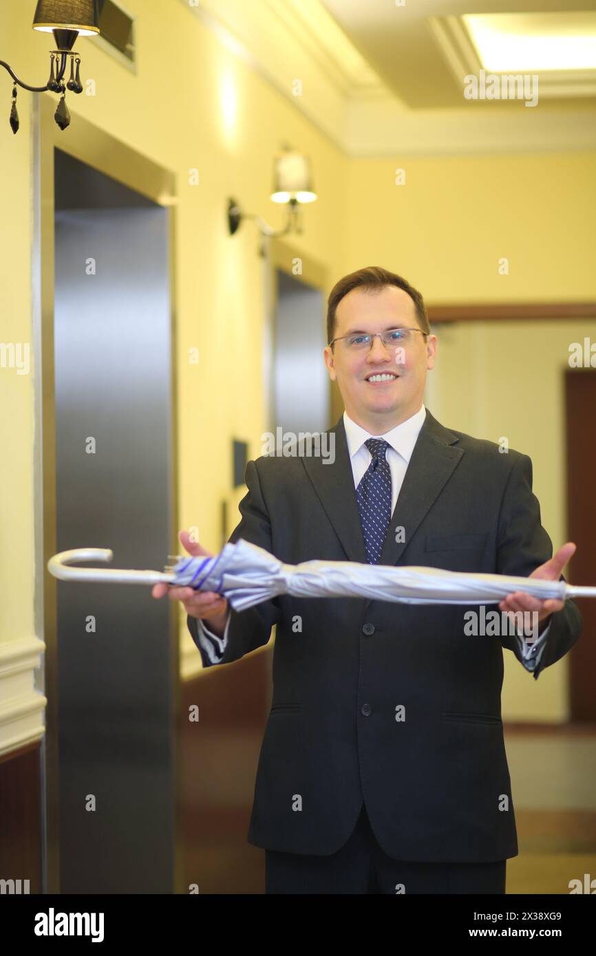 Un bell'uomo con una cravatta e una tuta da lavoro tiene un ombrello argentato nel corridoio vicino all'ascensore Foto Stock