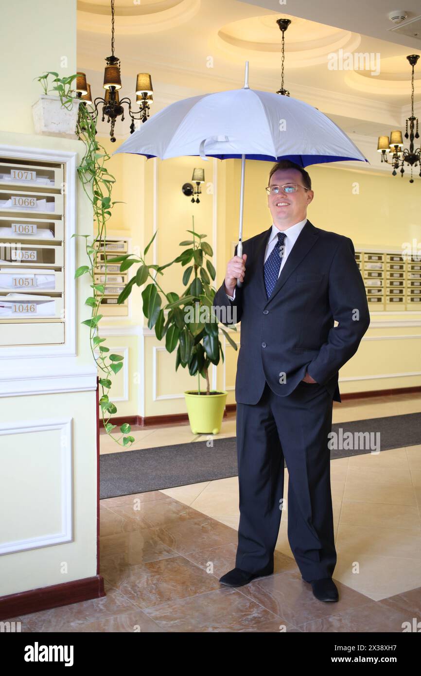 Bell'uomo con un abito da lavoro e cravatta in piedi con un ombrello aperto d'argento nel corridoio vicino alle cassette postali Foto Stock