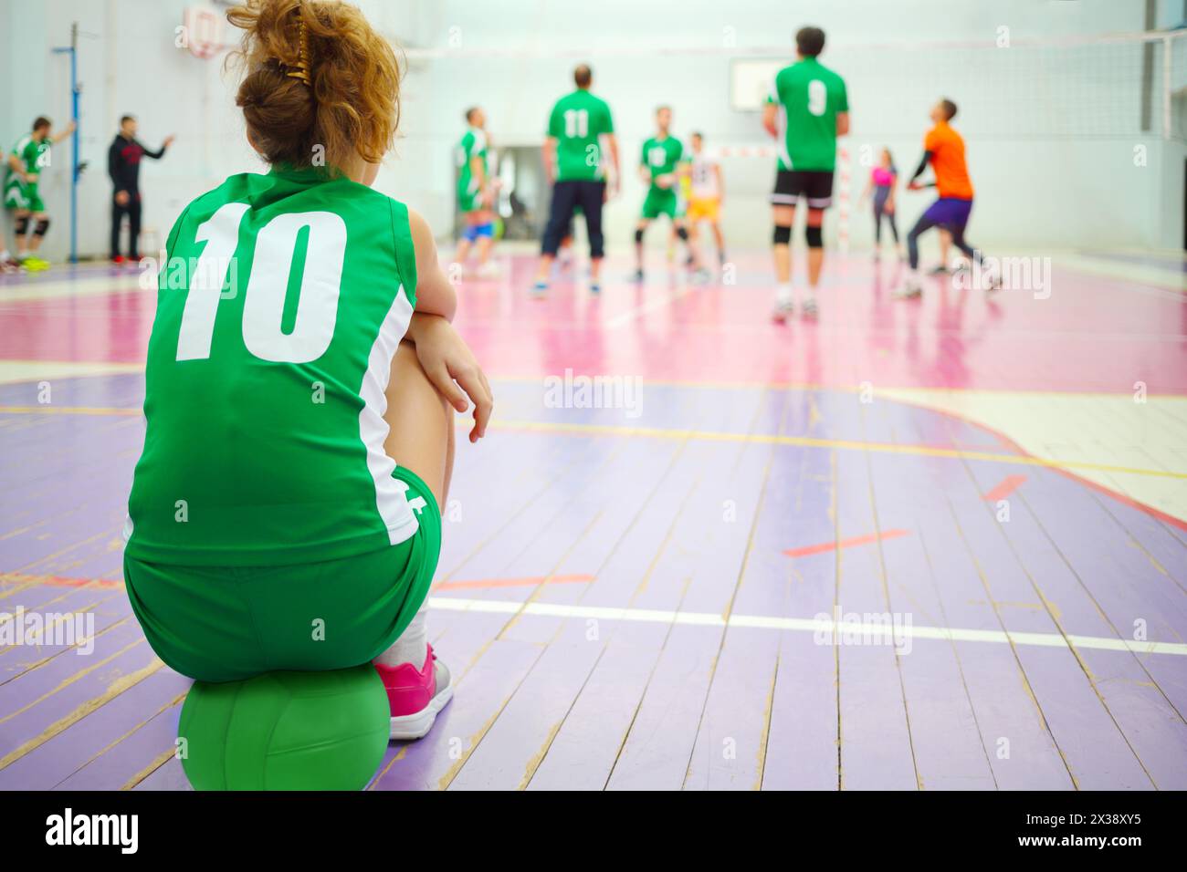 Una ragazza in verde si siede sulla palla in palestra durante la partita di pallavolo, vista posteriore, giocando a persone fuori fuoco Foto Stock