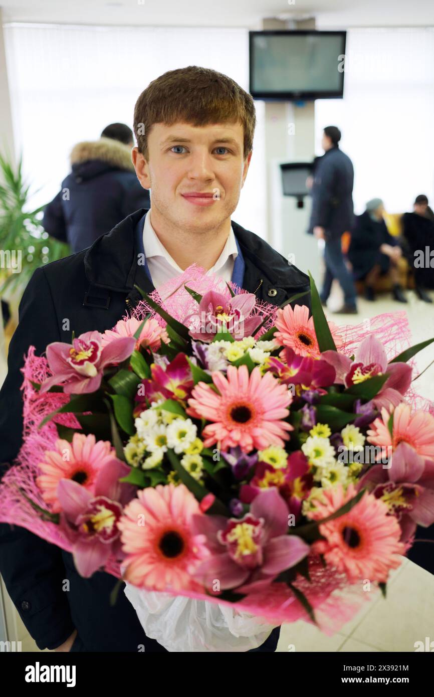 Felice giovane uomo tiene grande bouquet di fiori nella sala dell'ospedale di maternità, altre persone fuori fuoco Foto Stock