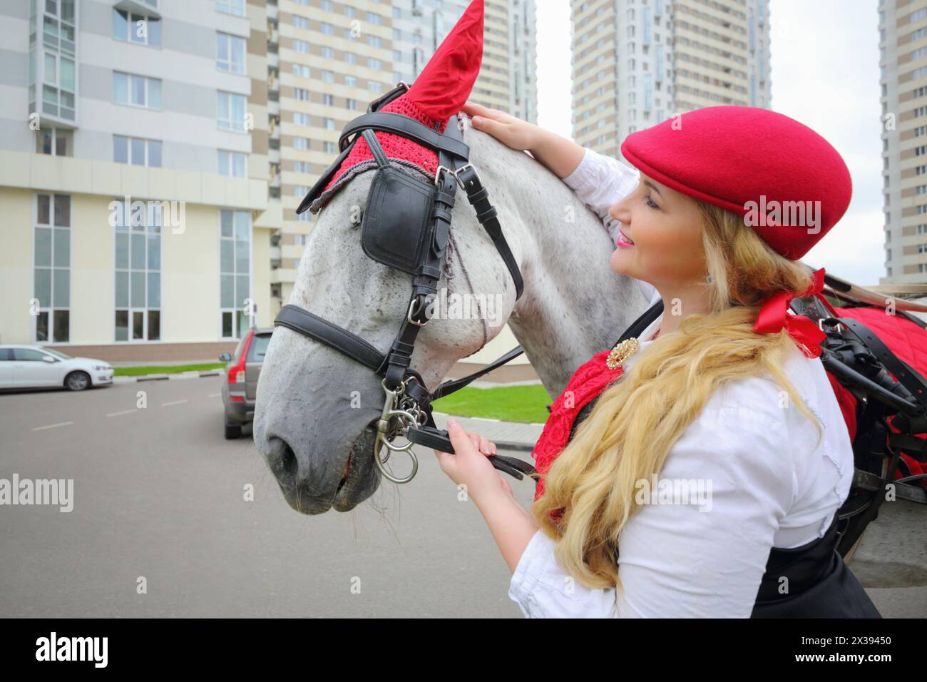 Coachman Woman sta con cavallo in un'imbracatura rossa vicino a edifici residenziali Foto Stock