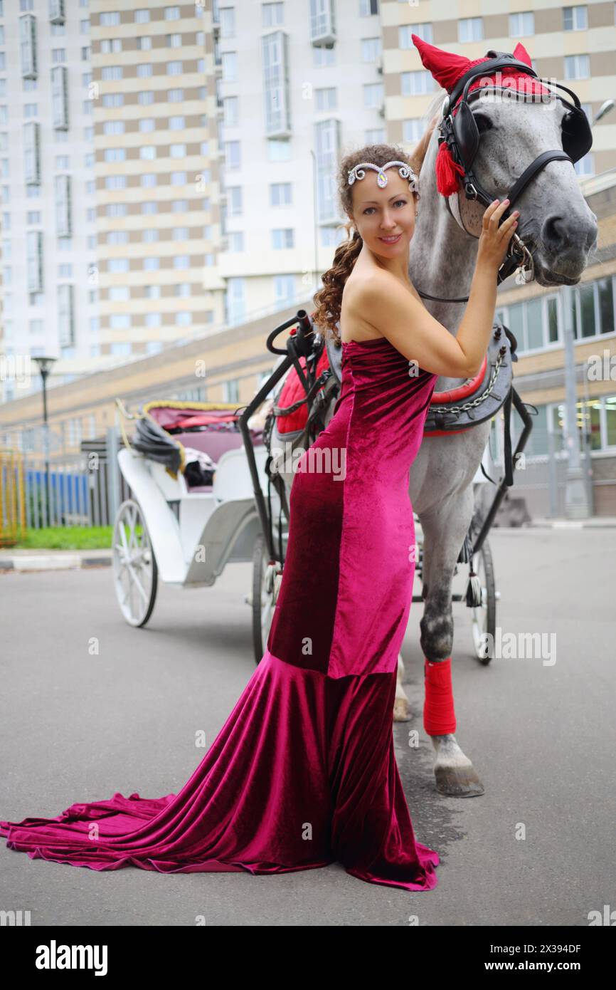 Una bella donna in abito rosso si trova vicino a un cavallo con un pullman vicino all'edificio residenziale Foto Stock
