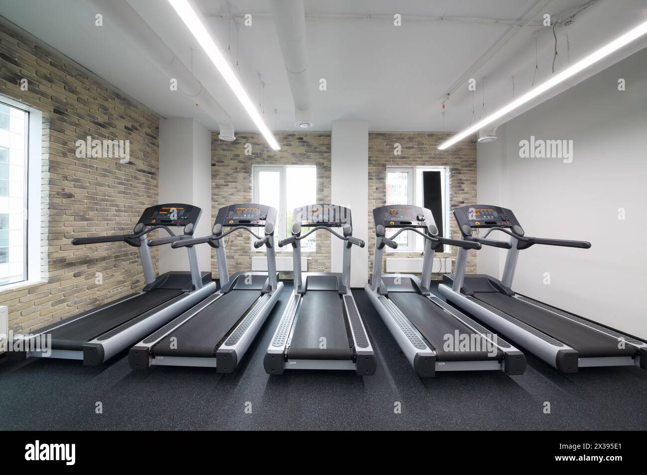 Cinque tapis roulant in una moderna sala vuota per il fitness con pareti in mattoni e finestre Foto Stock