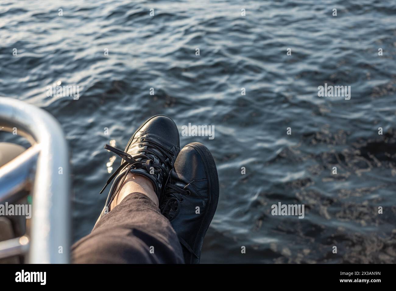 Piedi in sneakers nere appoggiati sul bordo di una barca, con il mare blu profondo sullo sfondo, che ritrae un momento marittimo rilassato. Foto di alta qualità Foto Stock