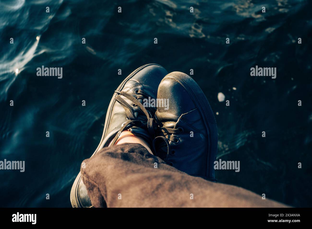 Persona con scarpe nere che penzolano i piedi sull'acqua, catturando un momento di calma e riflessione. Foto di alta qualità Foto Stock