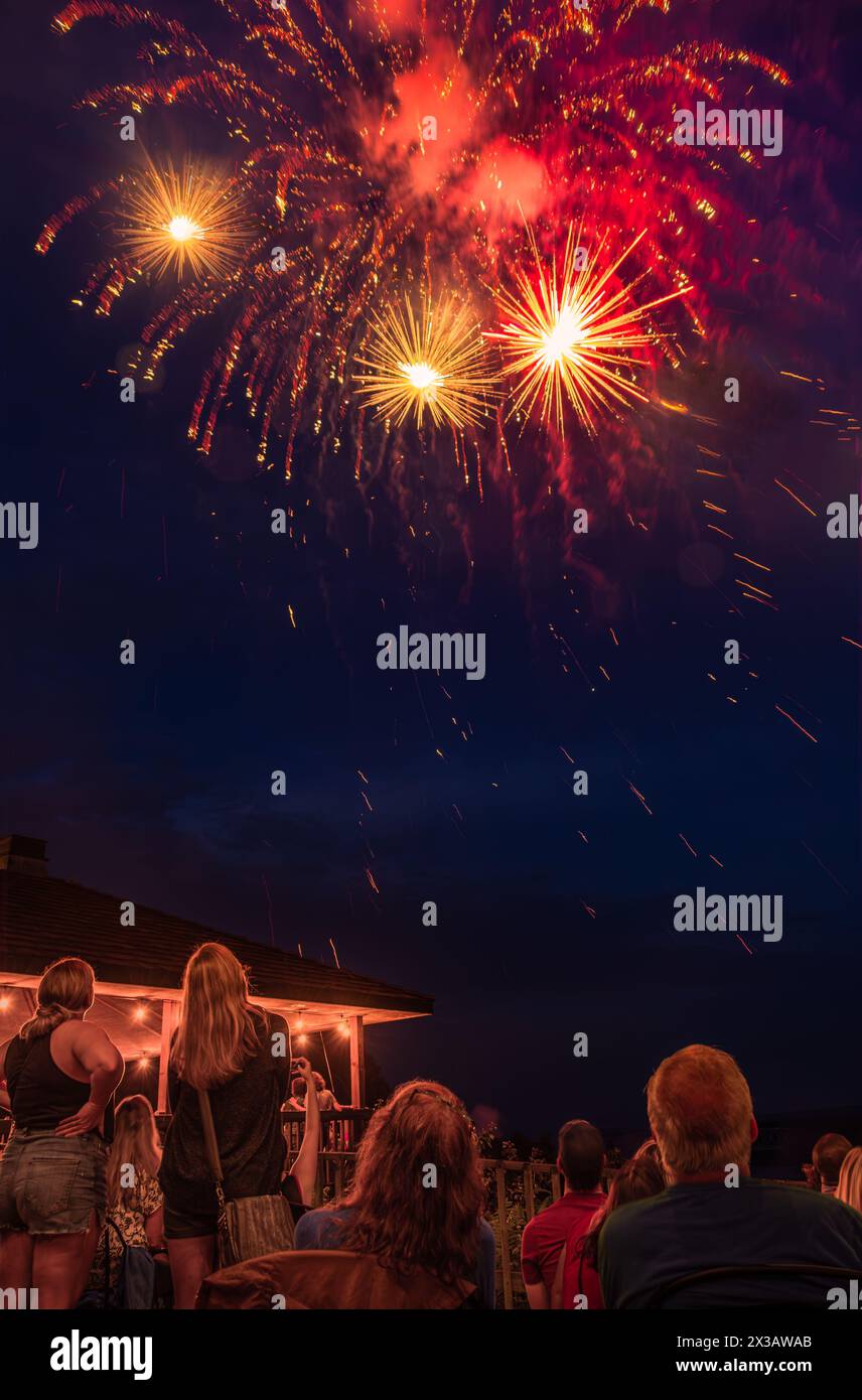 Fuochi d'artificio del 4 luglio nella piccola città del Midwest; figure di persone sedute e in piedi in primo piano; gazebo illuminato sullo sfondo Foto Stock