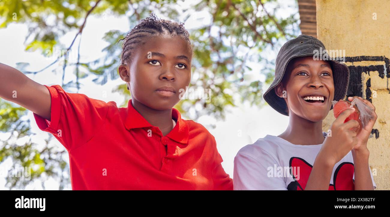 due ragazze africane del villaggio, che indossano un'acconciatura a treccia e un cappello, mangiano succo congelato da un sacchetto di plastica Foto Stock