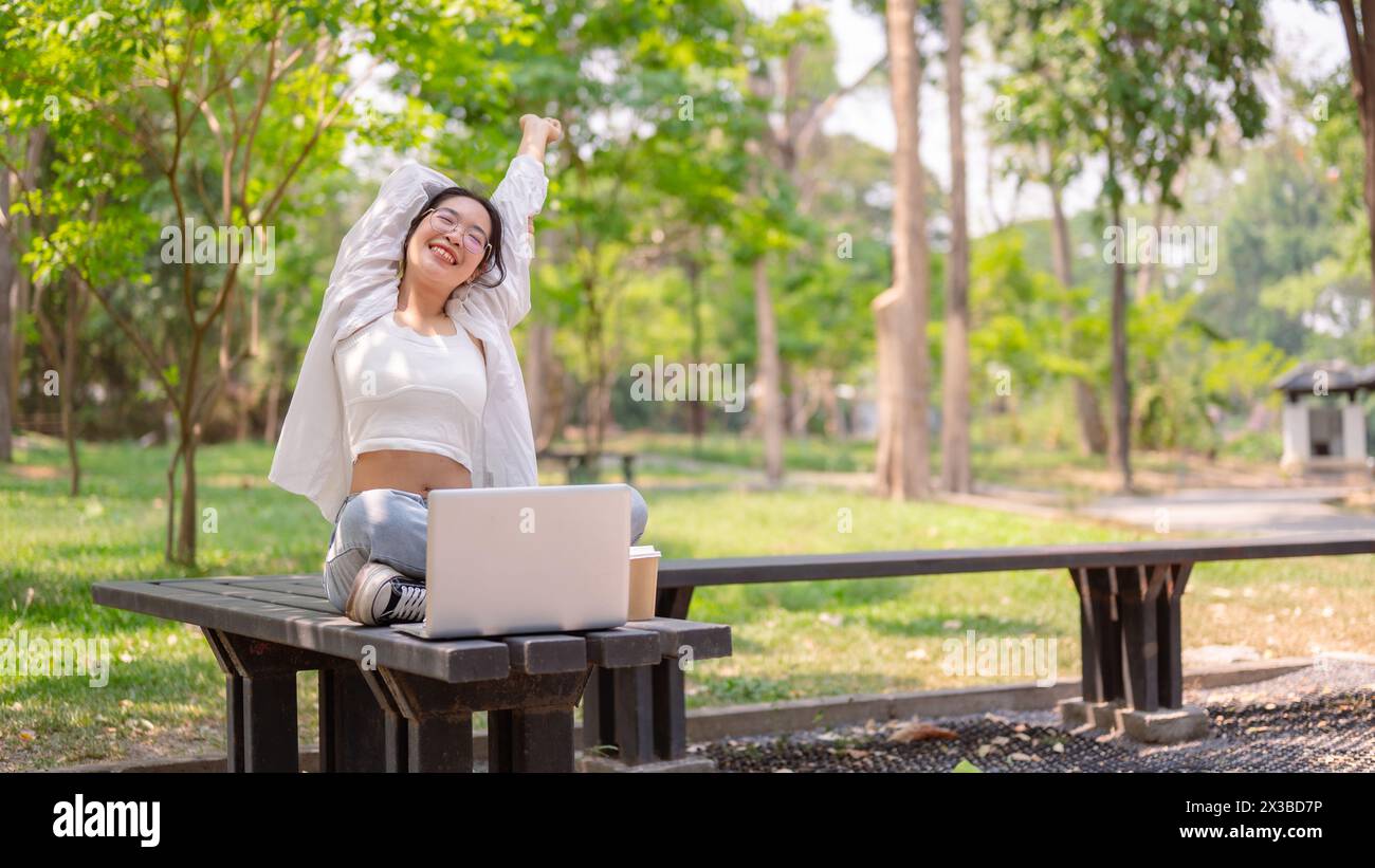 Una giovane donna asiatica spensierata e rilassata, in abbigliamento casual, siede su una panchina in un parco verde in una giornata luminosa, si piega indietro, si allunga le braccia e si gode la natura Foto Stock