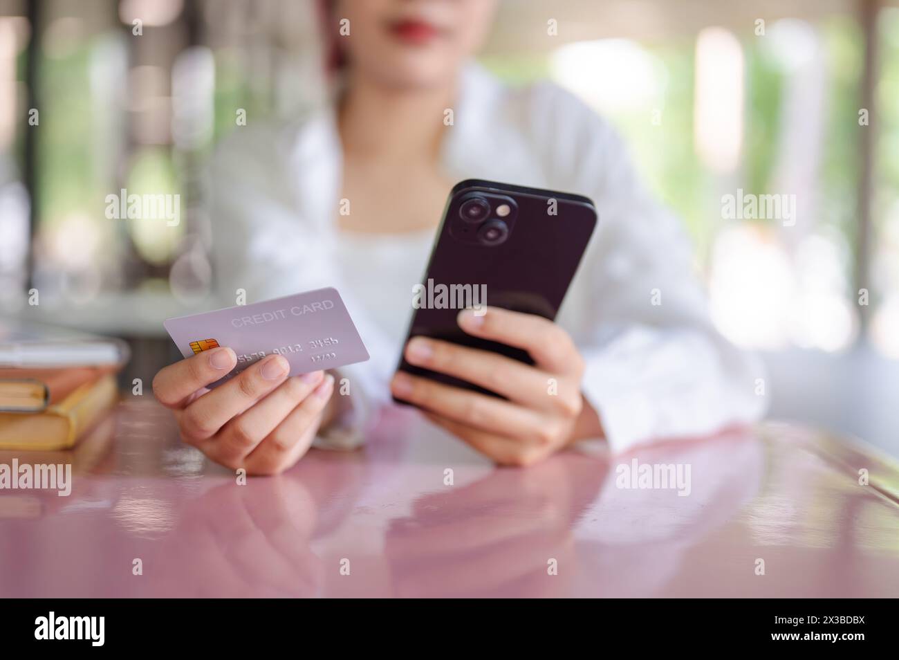 Un'immagine ritagliata di una studentessa asiatica in abbigliamento casual si siede a un tavolo in una caffetteria che tiene la sua carta di credito e lo smartphone, usando un mobil Foto Stock