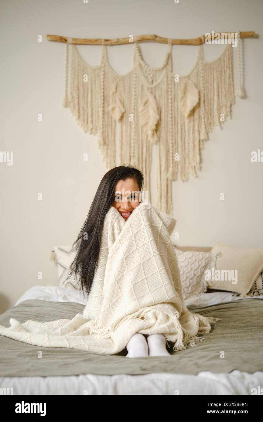 Donna sorridente seduta sul letto, avvolta in una morbida coperta Foto Stock