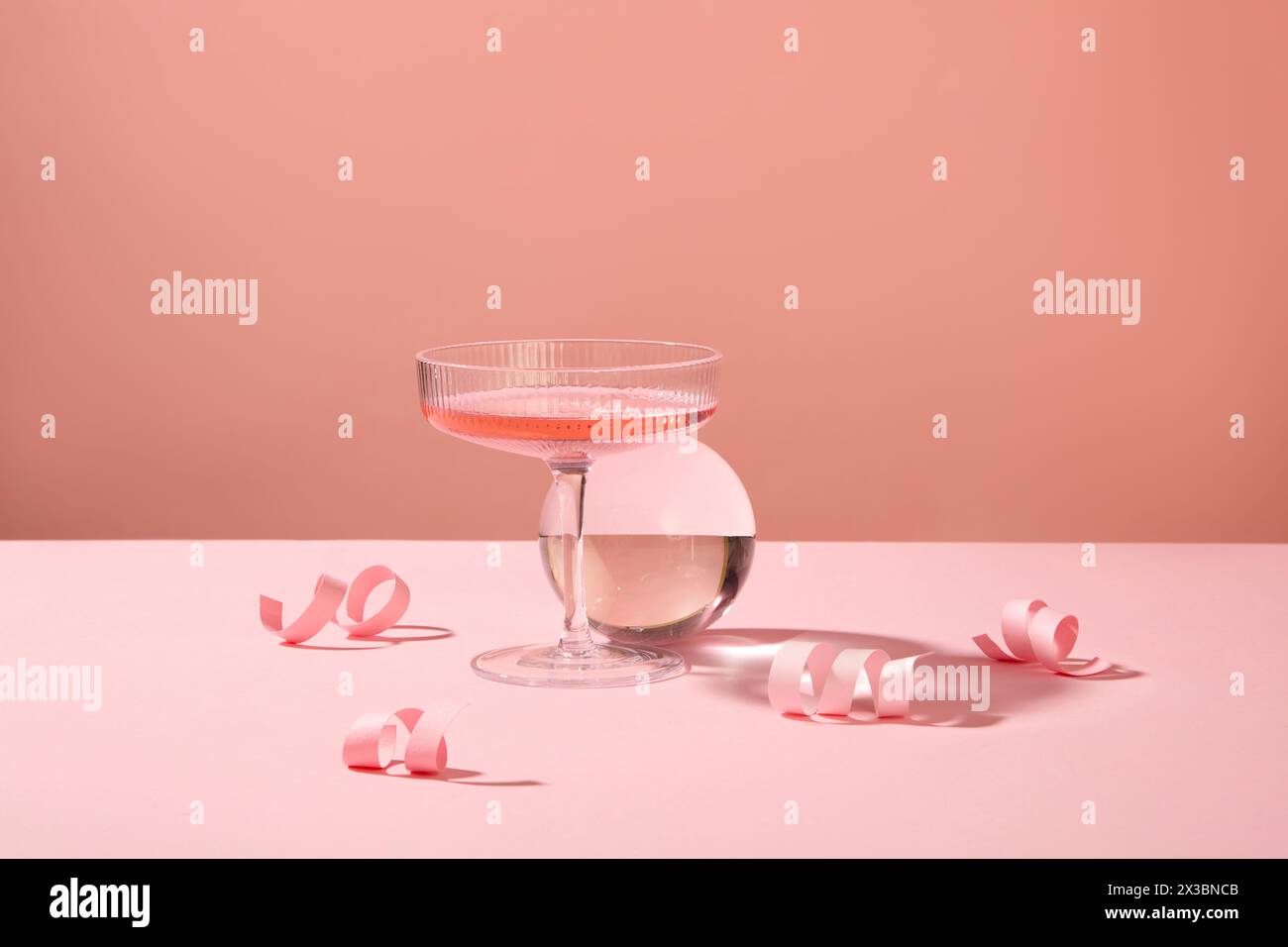 Scena minimale di un bicchiere dal design moderno pieno di vino rosa, decorato con una palla di vetro e alcuni fuochi d'artificio di carta rosa. Per il vostro saluto natalizio Foto Stock