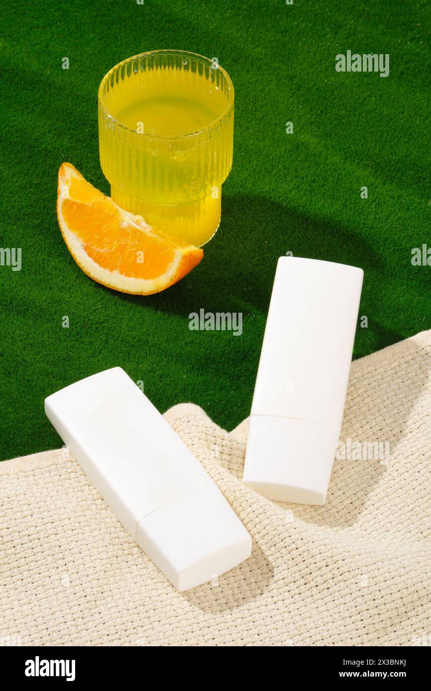 Scena minimale di due bottiglie di crema solare o crema idratante decorate sulla sciarpa. L'erba verde era composta da una tazza di succo d'arancia e una fetta d'arancia. Conta Foto Stock