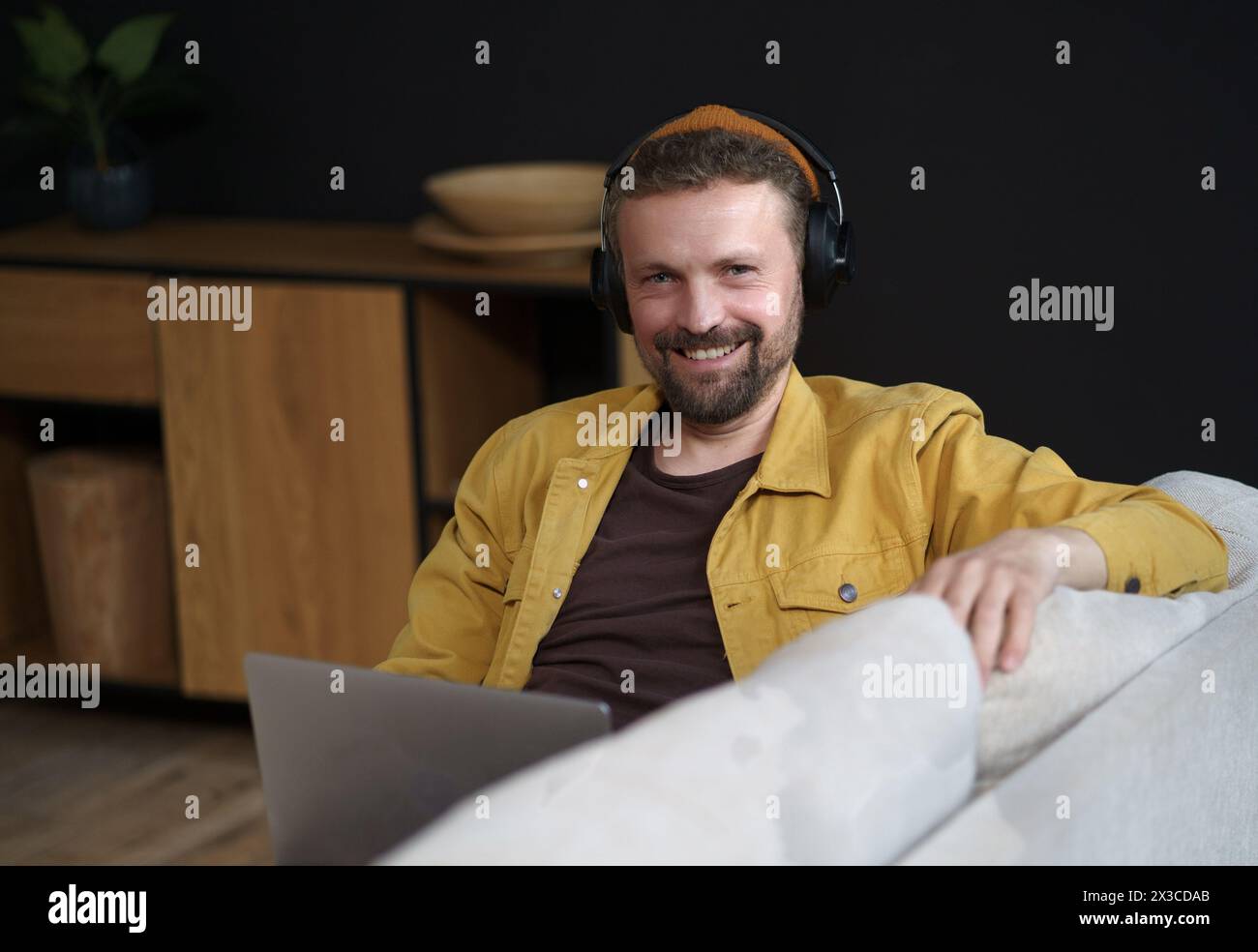 Un uomo che indossa le cuffie e una giacca gialla è seduto su un divano con un portatile davanti a sé. Sta sorridendo e si sta godendo il suo tempo Foto Stock