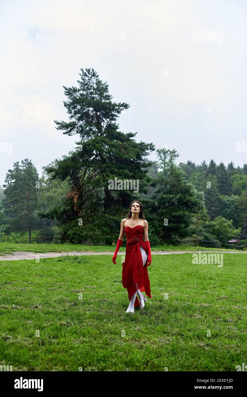Una giovane donna attraente con un abito rosso e guanti lunghi in piedi graziosamente in un campo, abbracciando la brezza estiva. Foto Stock