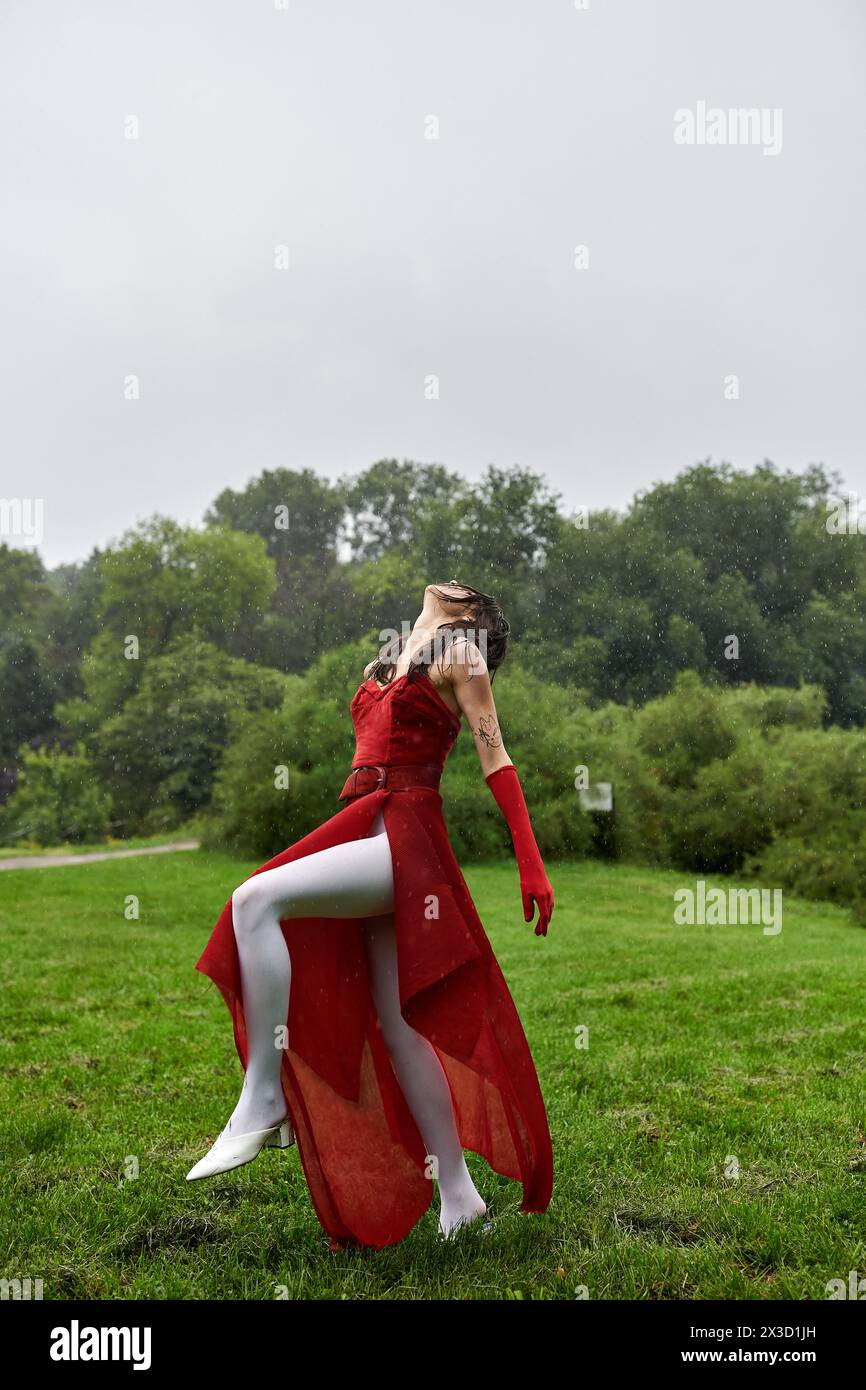 Una giovane donna elegante con un abito rosso fluente e lunghi guanti in piedi graziosamente in un lussureggiante campo verde. Foto Stock