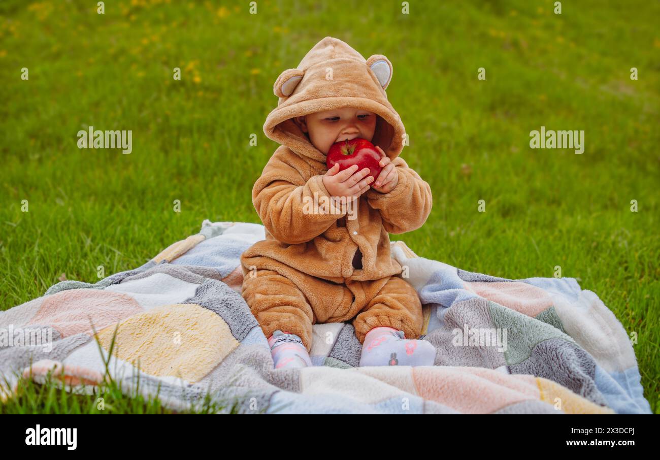 Una bambina con una tuta calda è seduta su un prato verde e mangia una mela Foto Stock