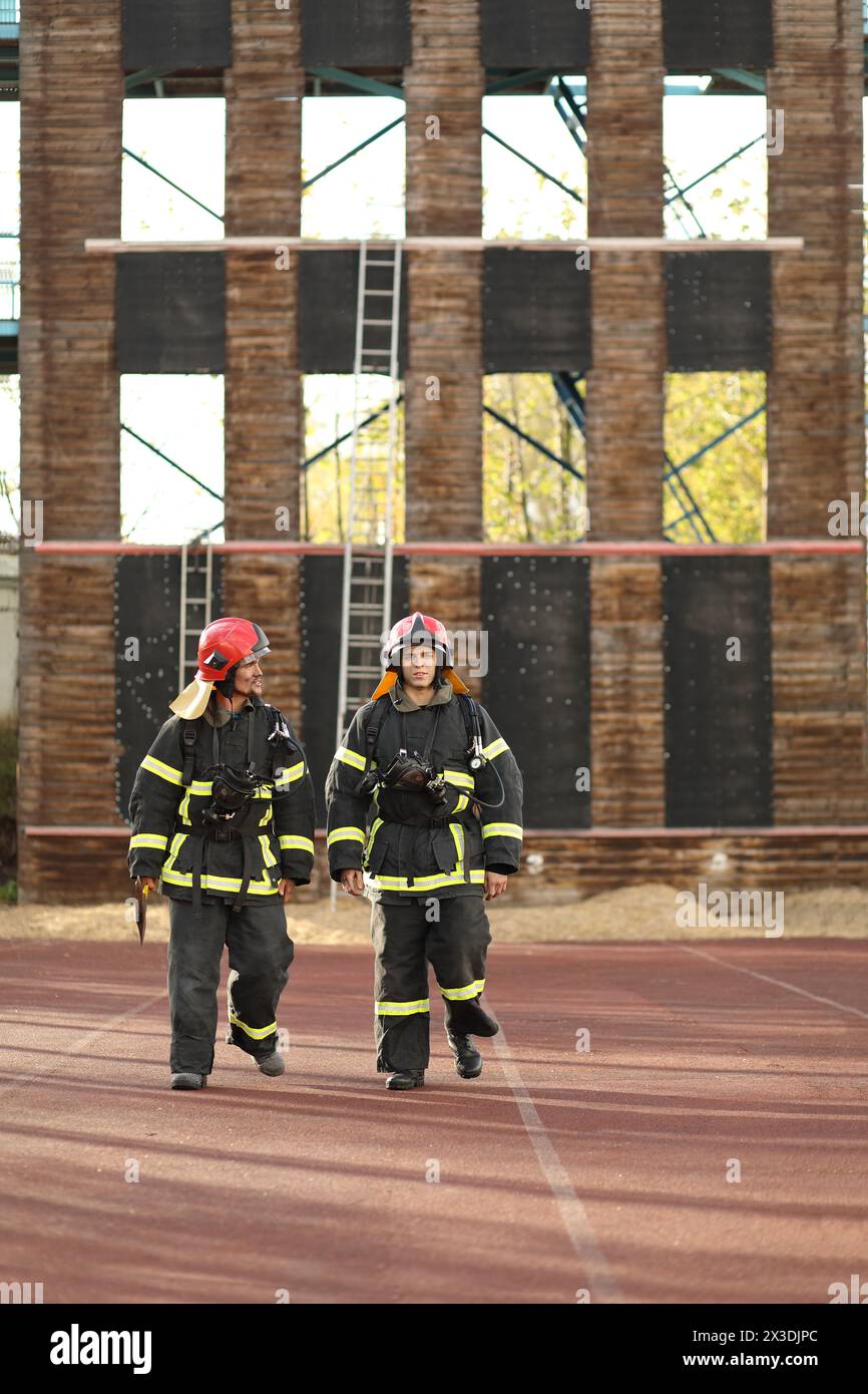 Due vigili del fuoco in equipaggiamento e caschi rossi nell'area di prova contro la torre antincendio Foto Stock