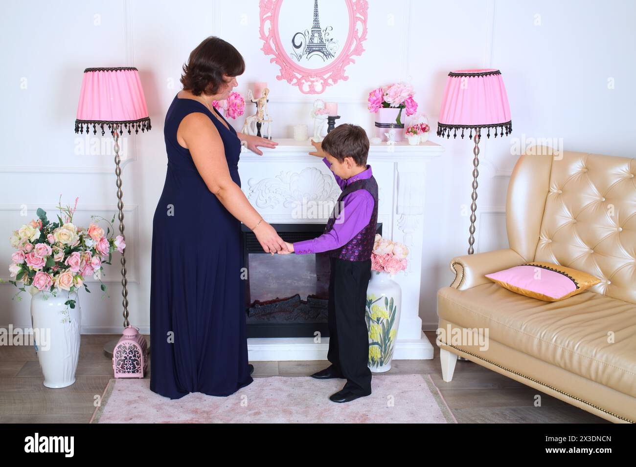 Madre e figlio in piedi vicino al caminetto nel soggiorno in uno stile romantico con due lampade da terra, divano e fiori Foto Stock