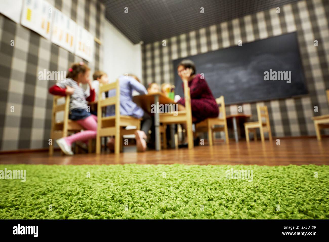 I bambini e l'insegnante di asilo nido siedono alle scrivanie in classe presso il club per bambini, vista ad angolo basso, focalizzazione sul rivestimento del pavimento, dof poco profondo. Foto Stock