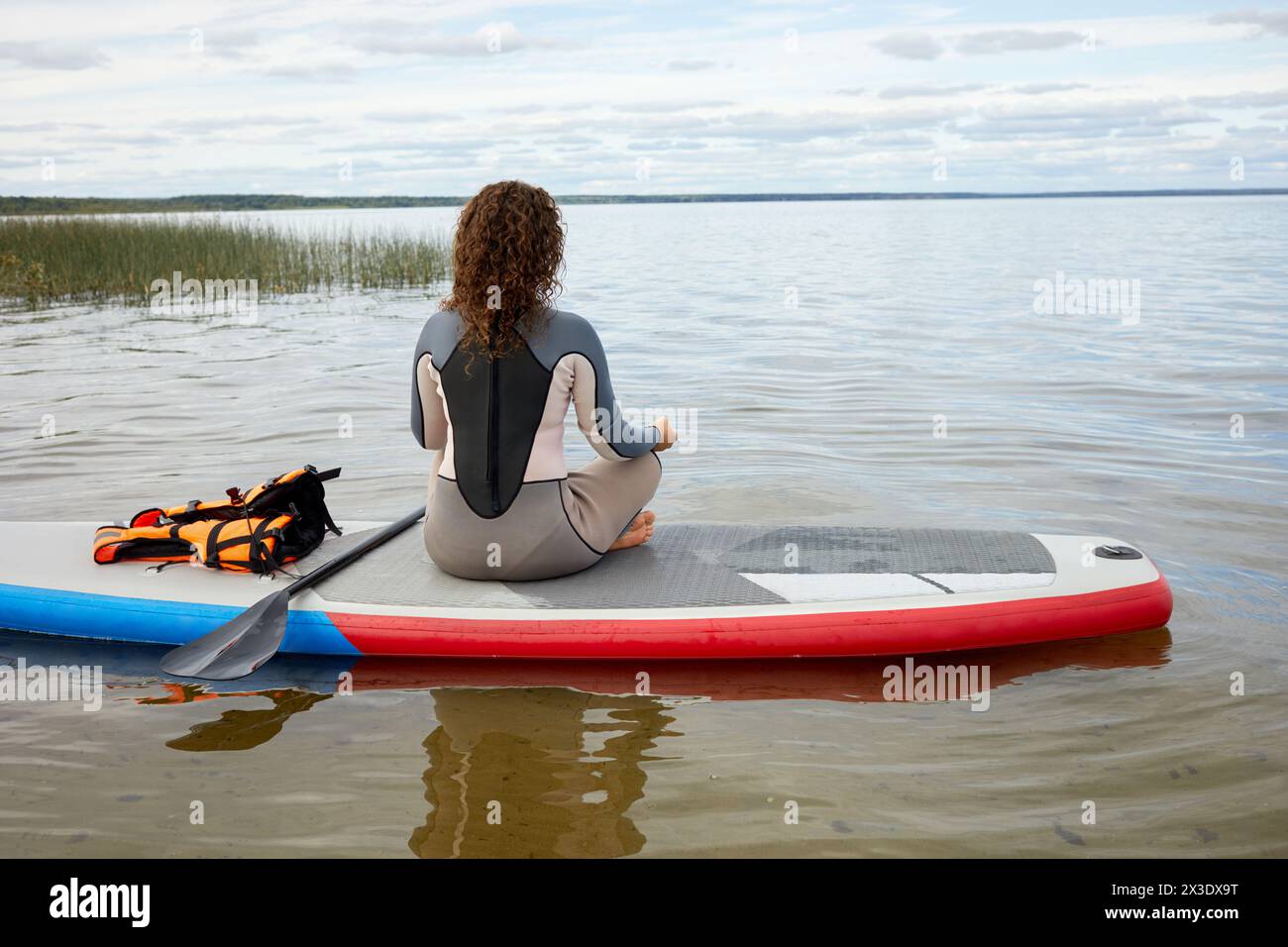 Donna con tuta rashguard seduto in posizione yoga sulla tavola SUP gonfiabile, vista posteriore. Foto Stock