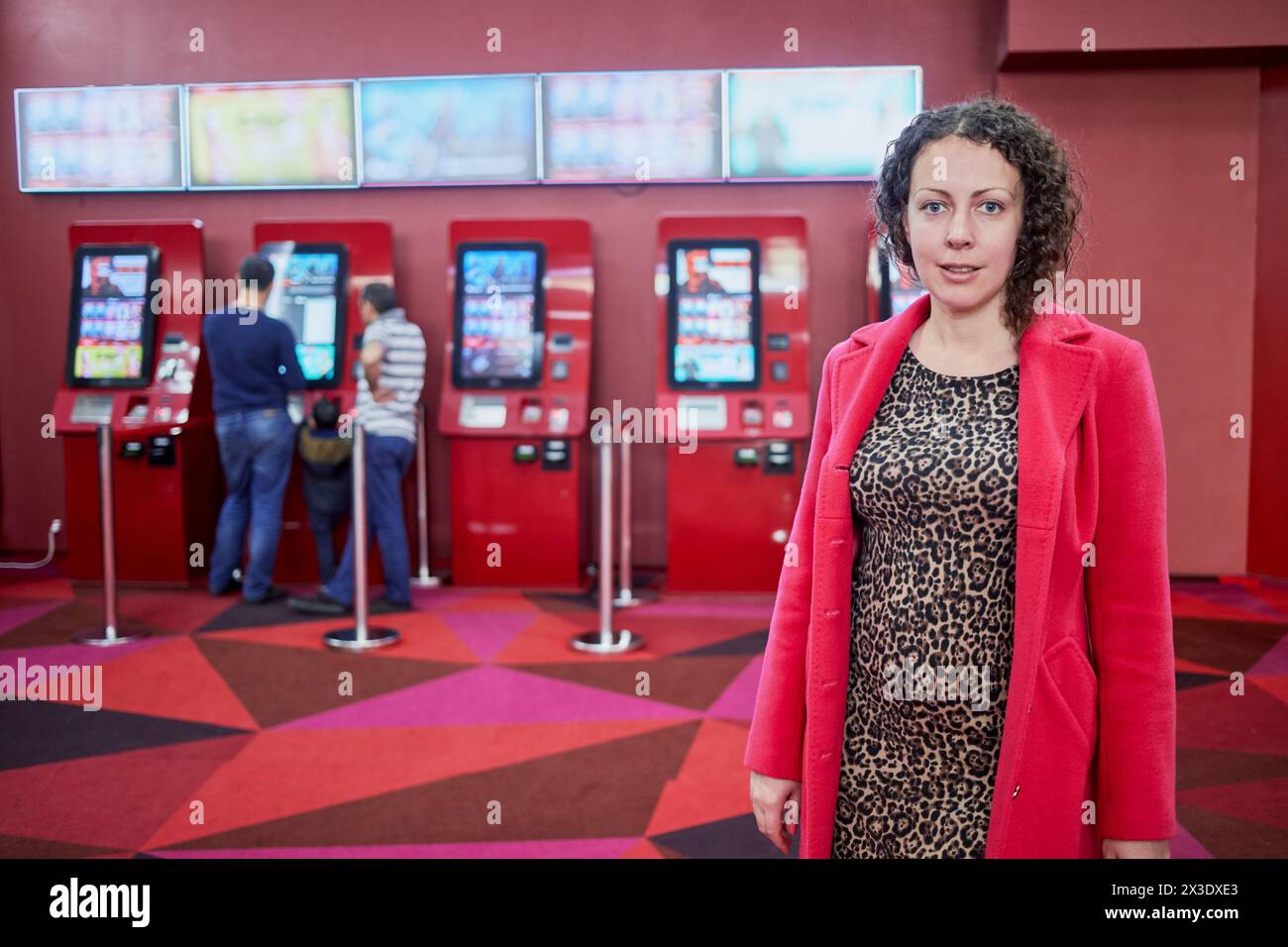 Una donna in camice rosso si trova al coperto in camera con distributori automatici di biglietti per il cinema. Foto Stock