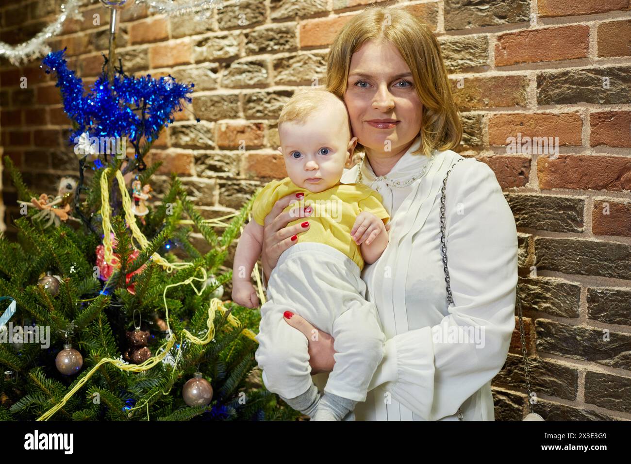 Donna sorridente con il bambino in braccio vicino all'albero di Natale contro la parete di mattoni rossi al coperto. Foto Stock