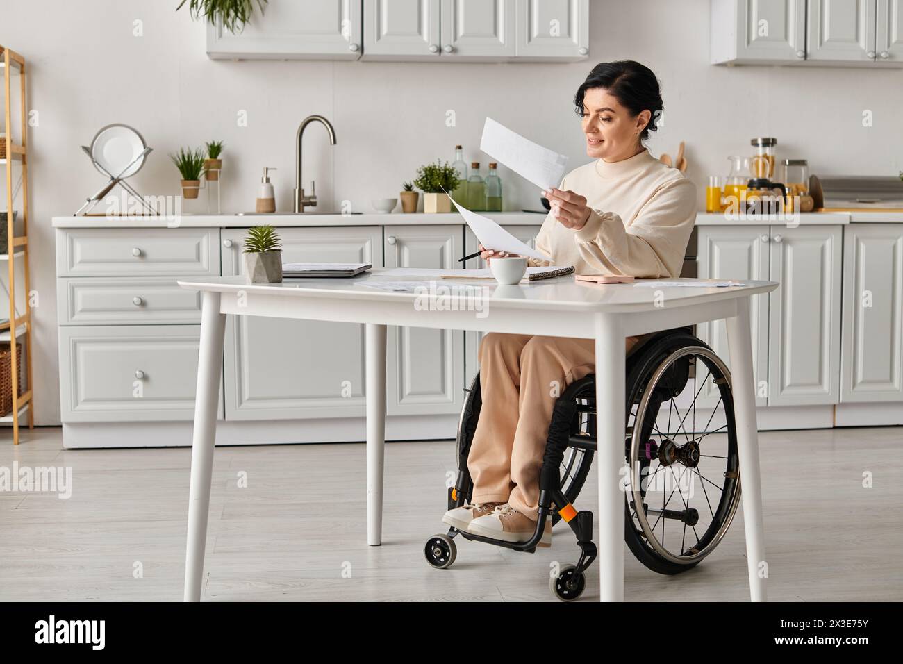 Una donna in sedia a rotelle siede nella sua cucina, profondamente pensata mentre tiene un pezzo di carta, concentrata sul suo lavoro creativo. Foto Stock