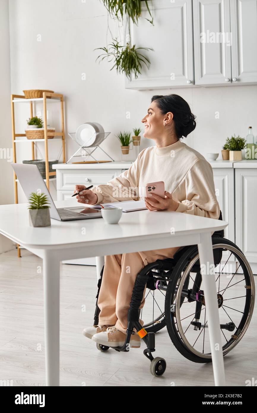 una donna disabile su una sedia a rotelle che lavora su un computer portatile nella sua cucina Foto Stock