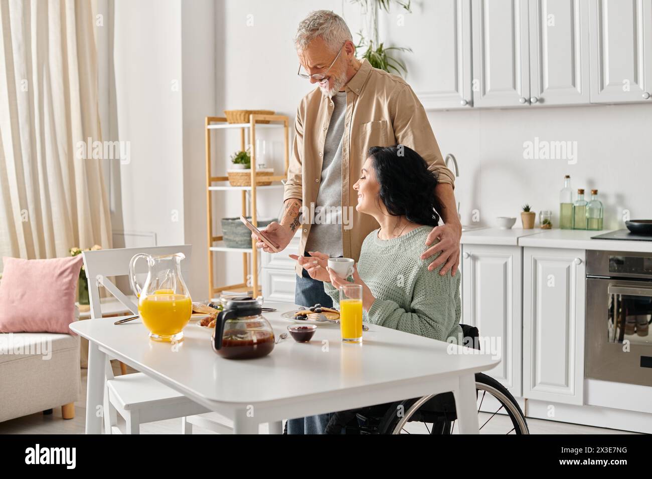 Un marito sta accanto alla moglie disabile su una sedia a rotelle, offrendo supporto e compagnia nella loro cucina di casa. Foto Stock