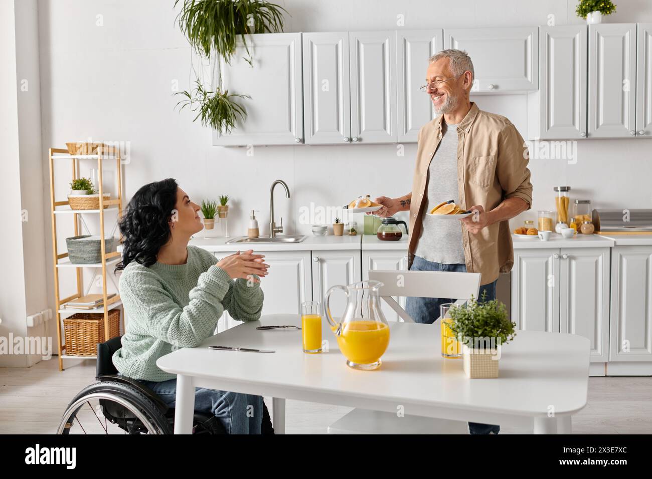 Una donna disabile in sedia a rotelle si impegna a conversare con il marito in un ambiente accogliente in cucina a casa. Foto Stock