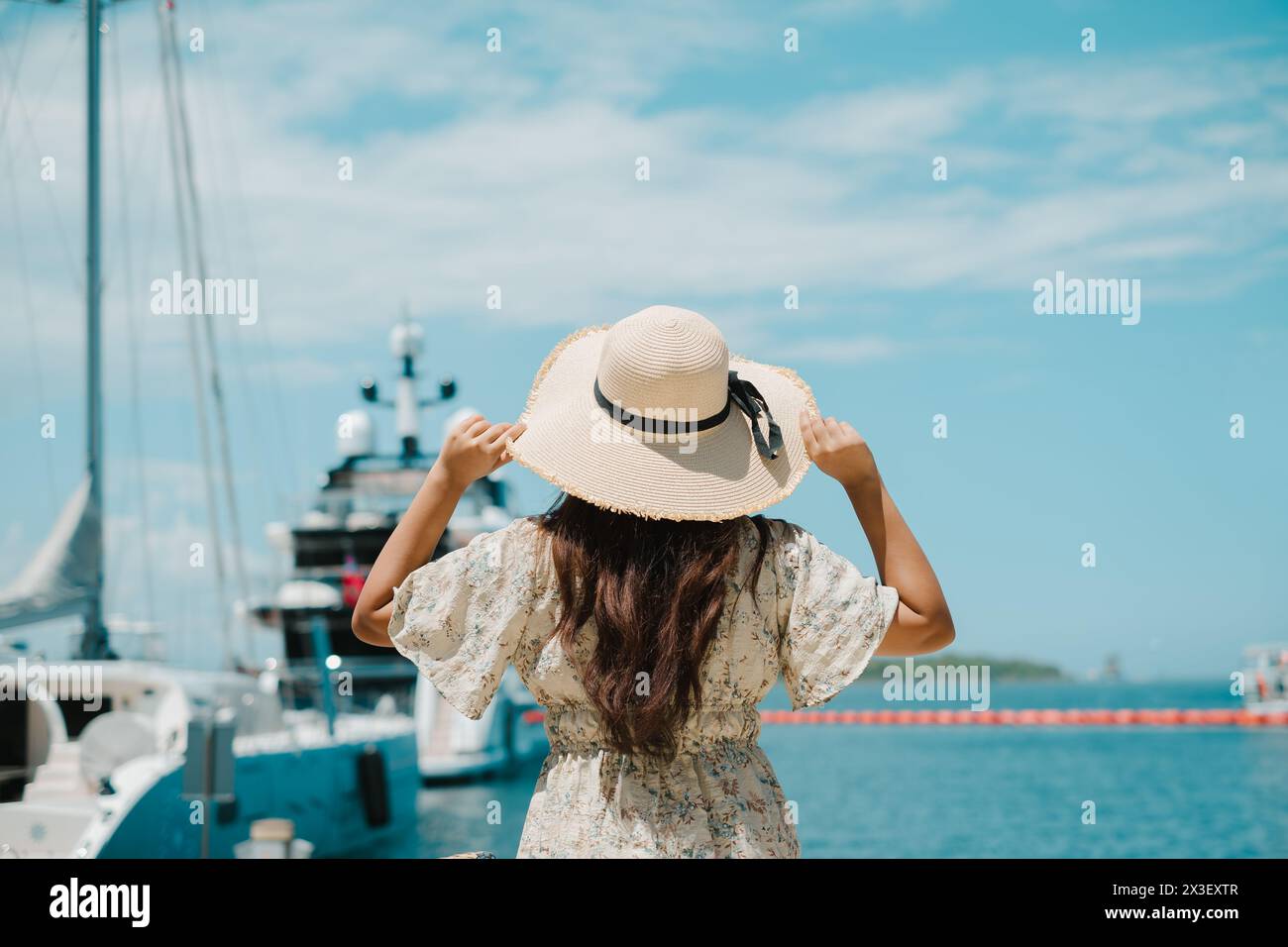 Giovane donna che tiene il suo cappello di paglia nel molo del porto degli yacht con una barca sfocata. Sfondo bokeh Foto Stock