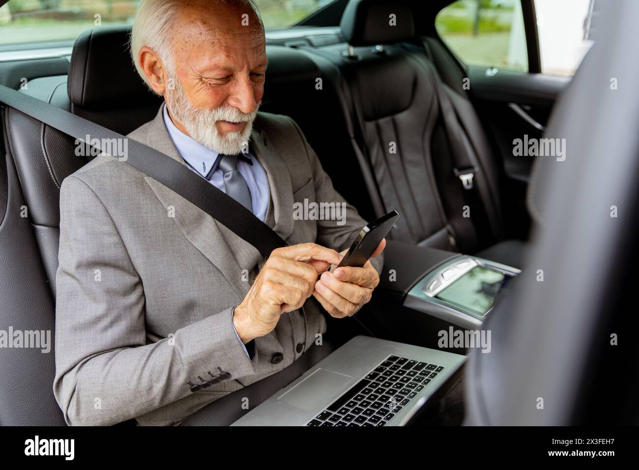 Un signore anziano in tuta esamina i messaggi sul suo cellulare durante un viaggio in auto, il suo portatile nelle vicinanze Foto Stock