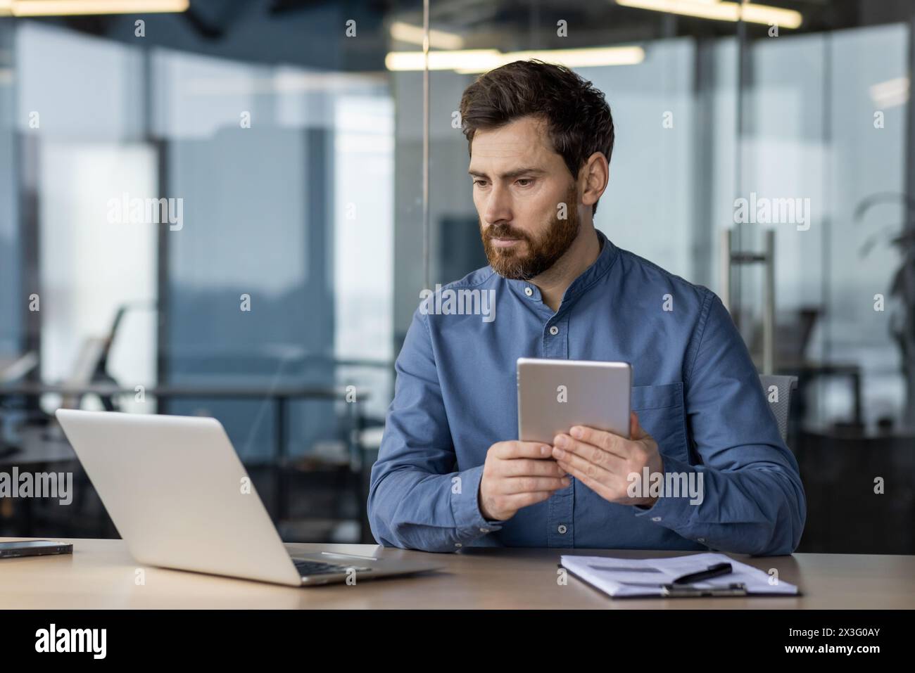 Un giovane serio lavora in ufficio a una scrivania, ha una coperta e guarda con attenzione lo schermo di un notebook, confronta i dati ed effettua un'analisi. Foto Stock