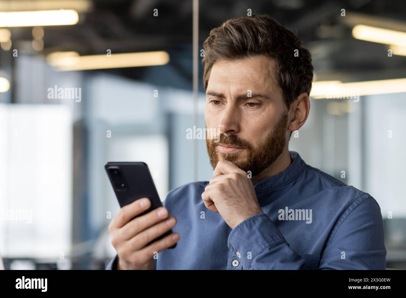 Foto ravvicinata di un giovane serio e premuroso che è in ufficio e legge un messaggio, guarda lo schermo del telefono cellulare, tiene la mano per la barba. Foto Stock