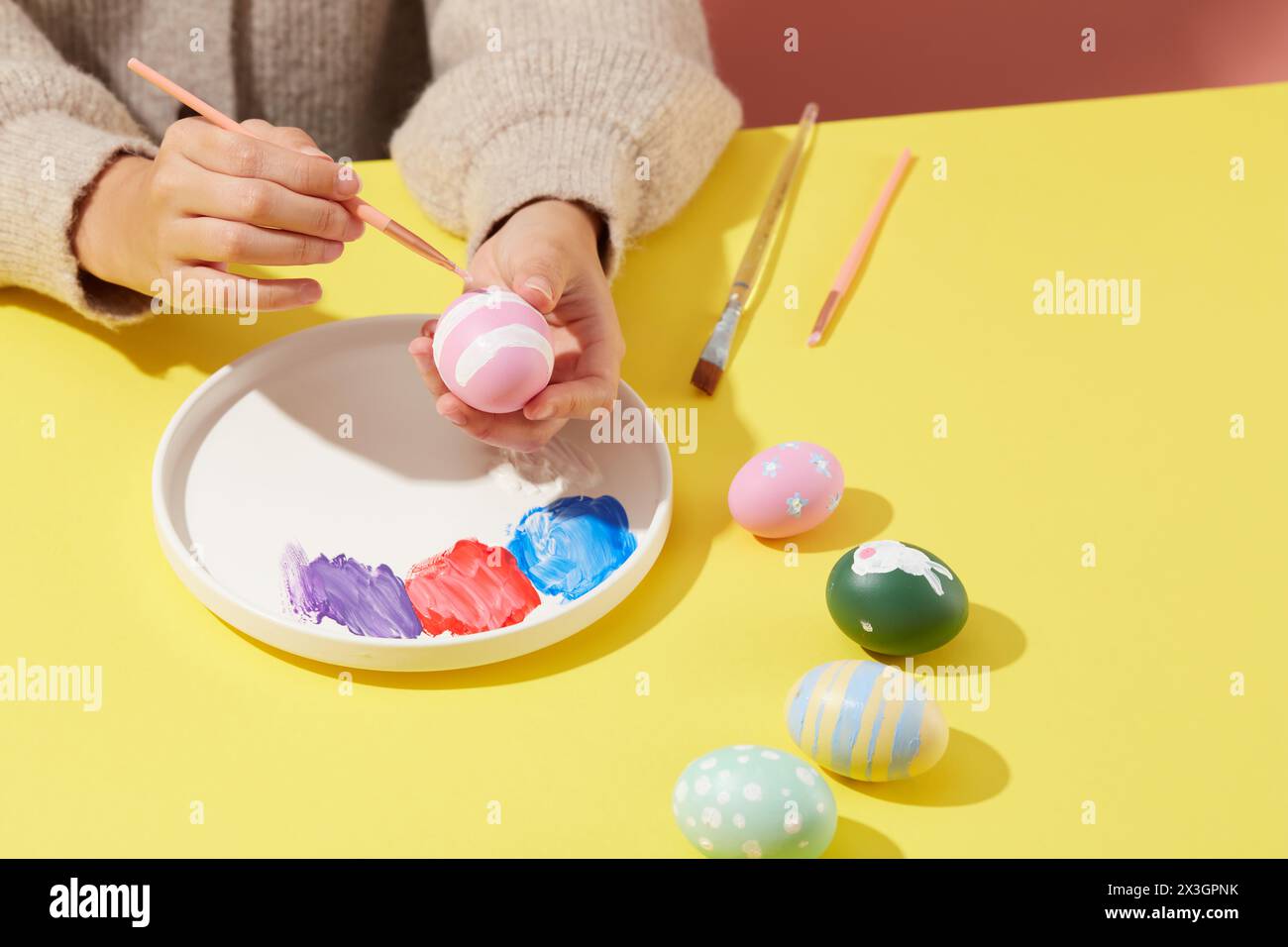 Una donna che indossa un maglione dipinge un uovo di Pasqua con un pennello, sul piatto caratterizzato da tre colori di vernice. Alcune uova di Pasqua con diverse patte Foto Stock