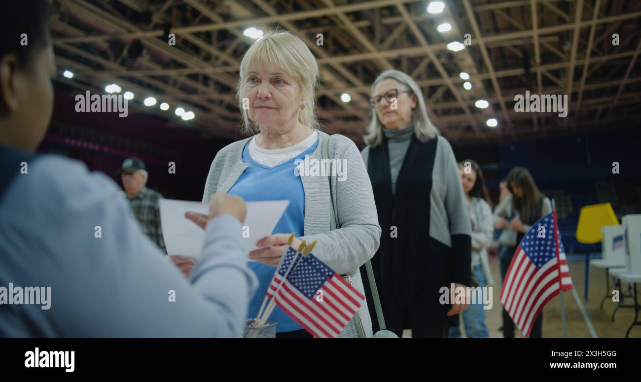 Una donna anziana parla con un funzionario e prende il bollettino. I cittadini americani vengono a votare al posto elettorale. Il giorno delle elezioni nazionali negli Stati Uniti. Razze politiche di candidati presidenziali statunitensi. Foto Stock